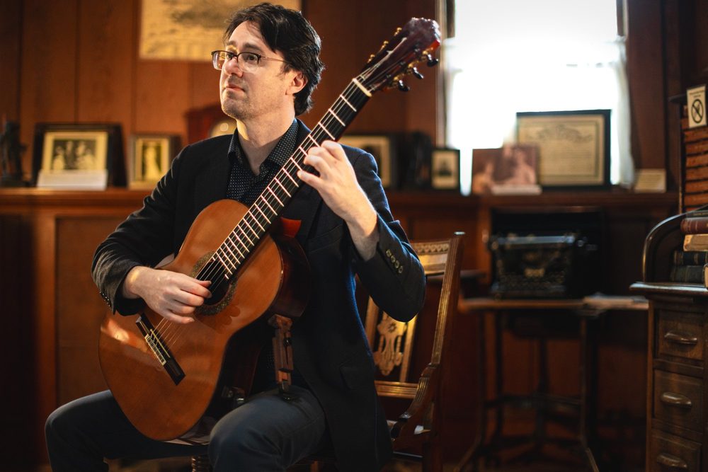Yuri Liberzon with his guitar, looking up, in a wood-panelled room